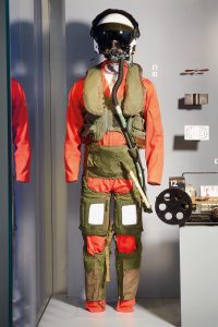A full-body display of a jet pilot flight suit consisting of an orange jumpsuit with green anti-G suit components, a green life vest, and a white flight helmet with a dark visor and attached oxygen mask.