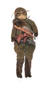 Small figure of a female aviator. Flying suit and helmet are green velvet with a beige face, on which are drawn female features. The figure wears brown boots, a fur collar and two sashes. The upper sash is blue with gold text reading 'London-Cape' whilst the lower is in red with gold text reading 'London-India'. A pair of woolen googles are also part of the item but have become detached.