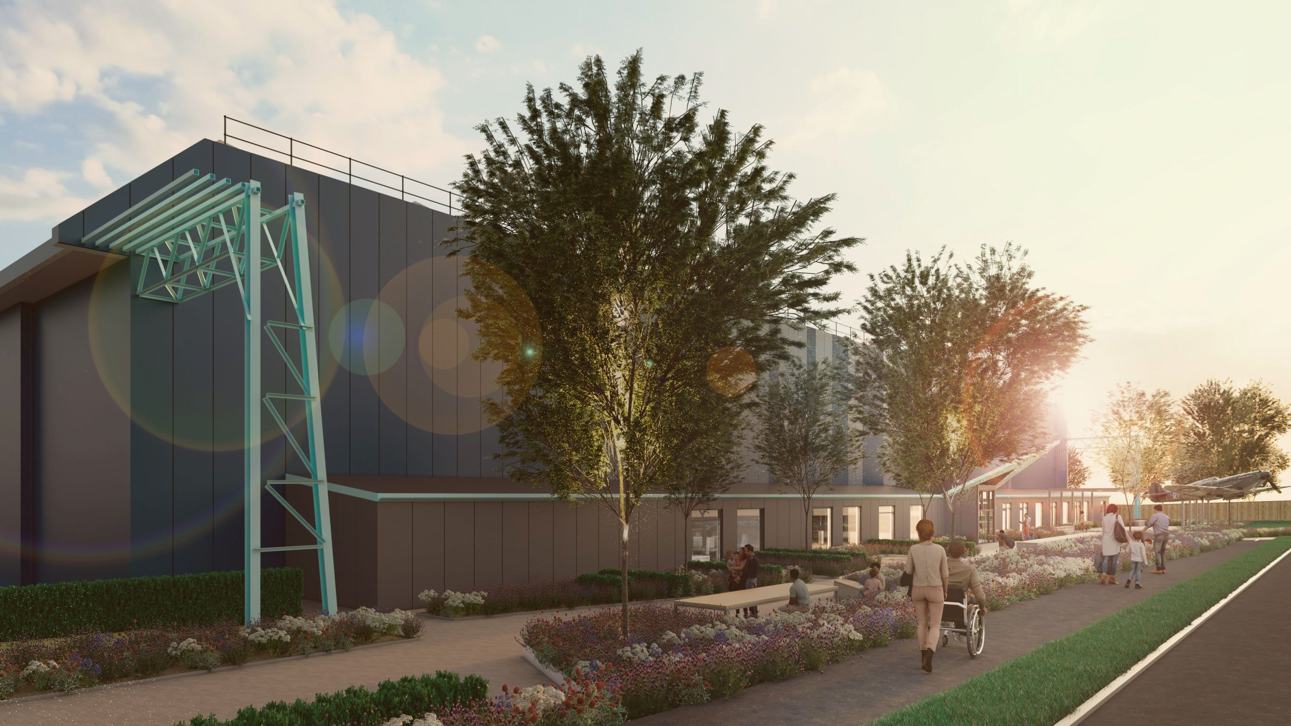 The exterior of an aircraft hangar with trees and plants outside while people walk along the front towards the sunset.