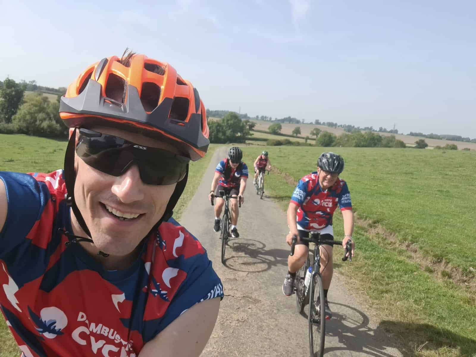 A white man wearing an orange bike helmet smiles as he takes a selfie of himself and three other cyclists behind him