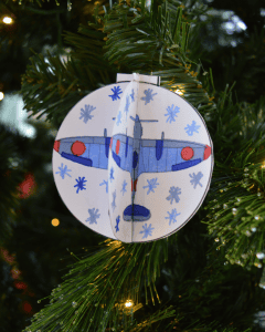 A hand made paper bauble hanging on a Christmas tree. There is a Second World War style plane printed on the bauble which has been coloured in by hand and snowflakes are doodled around it.