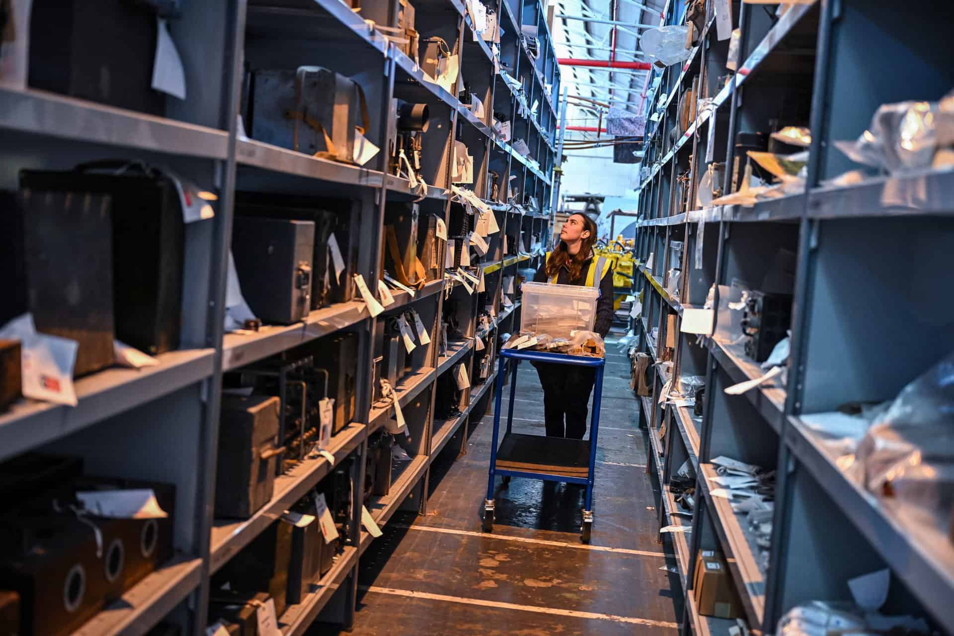 A member of the stafford staff team walking down an aisle with tall racking either side. There is a trolley she is pushing with a box on top.