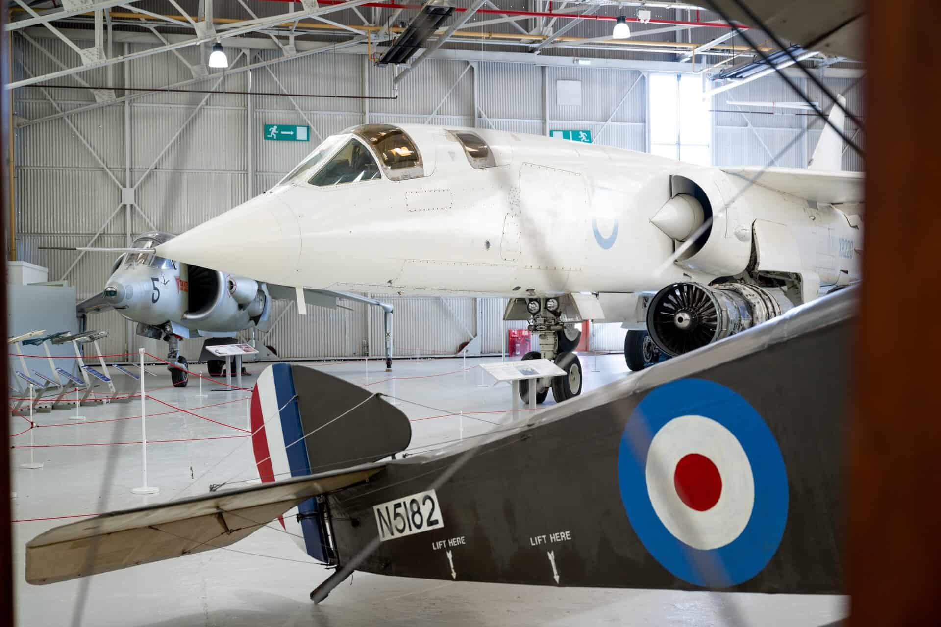 A white jet aircraft displayed inside an aviation museum hangar, viewed from the side, with its nose pointed left and landing gear down. In the foreground, the tail and wing of an older RAF aircraft with a blue, white, and red roundel partially frame the scene. Overhead lights, metal beams, and information placards surround the aircraft