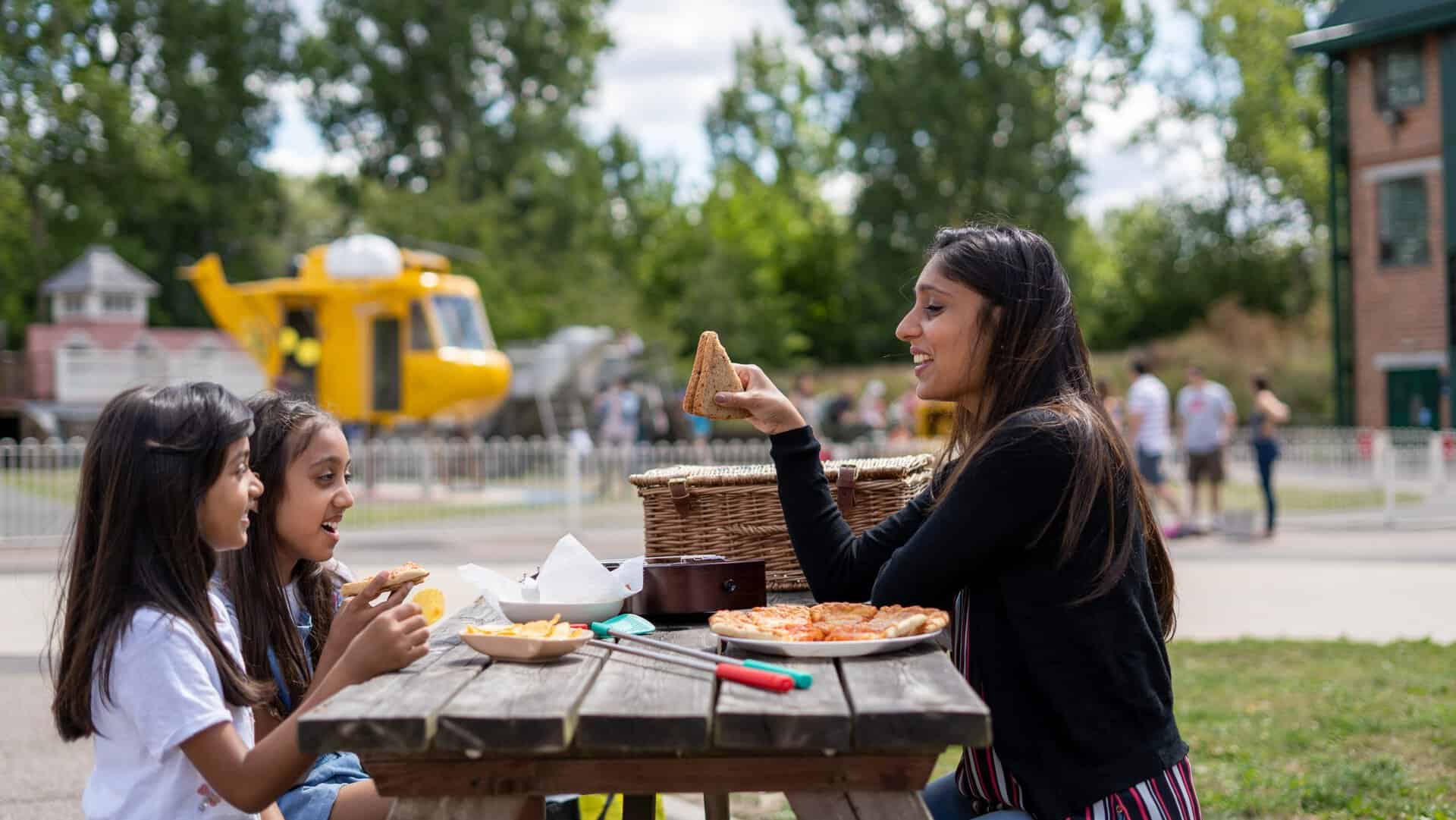 A woman and two children enjoying a picnic outdoors at the RAF Museum, with a helicopter in the back.